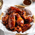 Homemade crispy hoisin chicken wings coated in sticky sweet Asian glaze, topped with sesame seeds, served in bamboo bowl with dipping sauce and chillies on light background